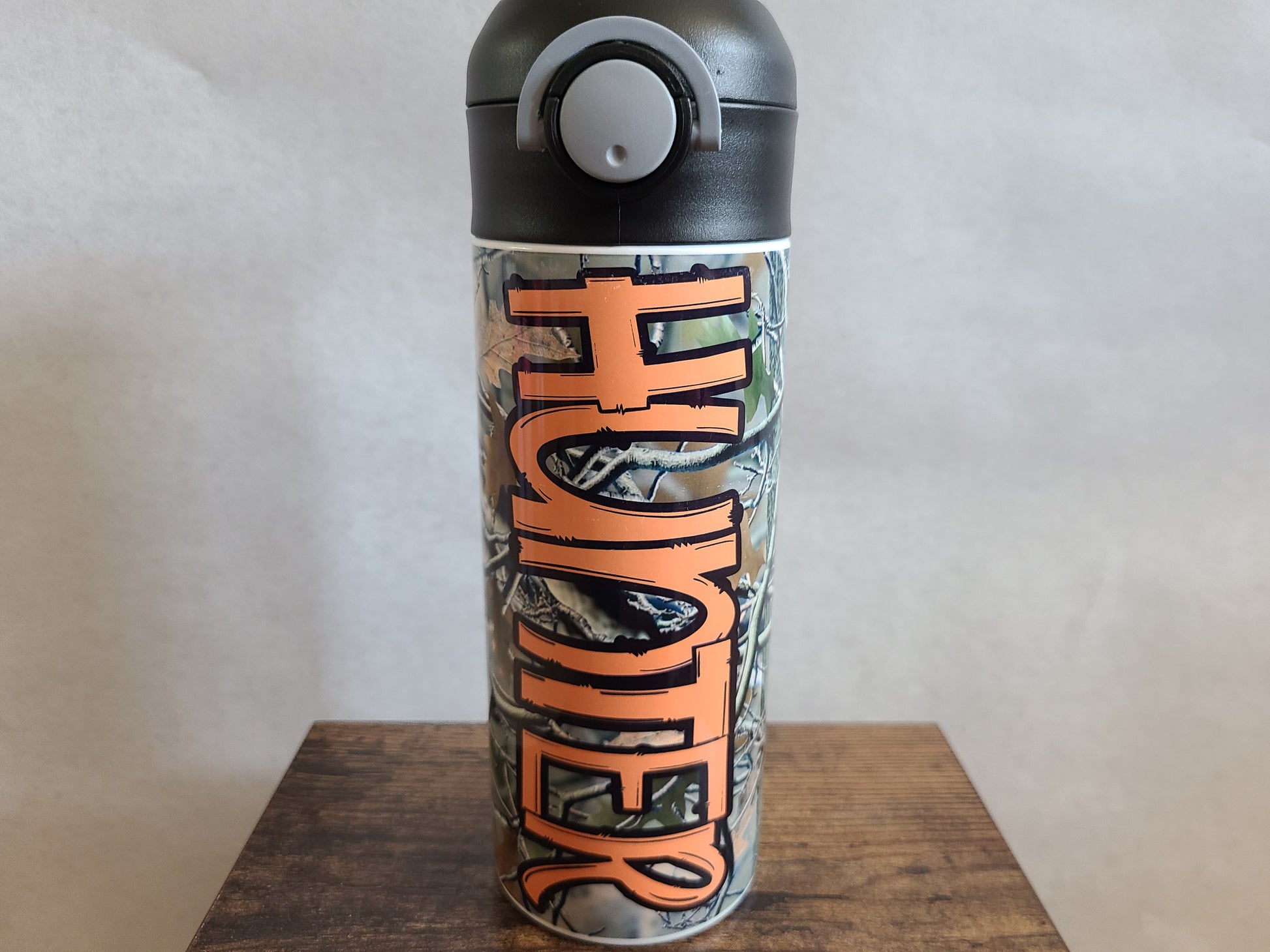 12 oz water bottle with a black lid that has a built-in straw, locking push button and handle. This design features real tree camo with orange personalization. Back to School.