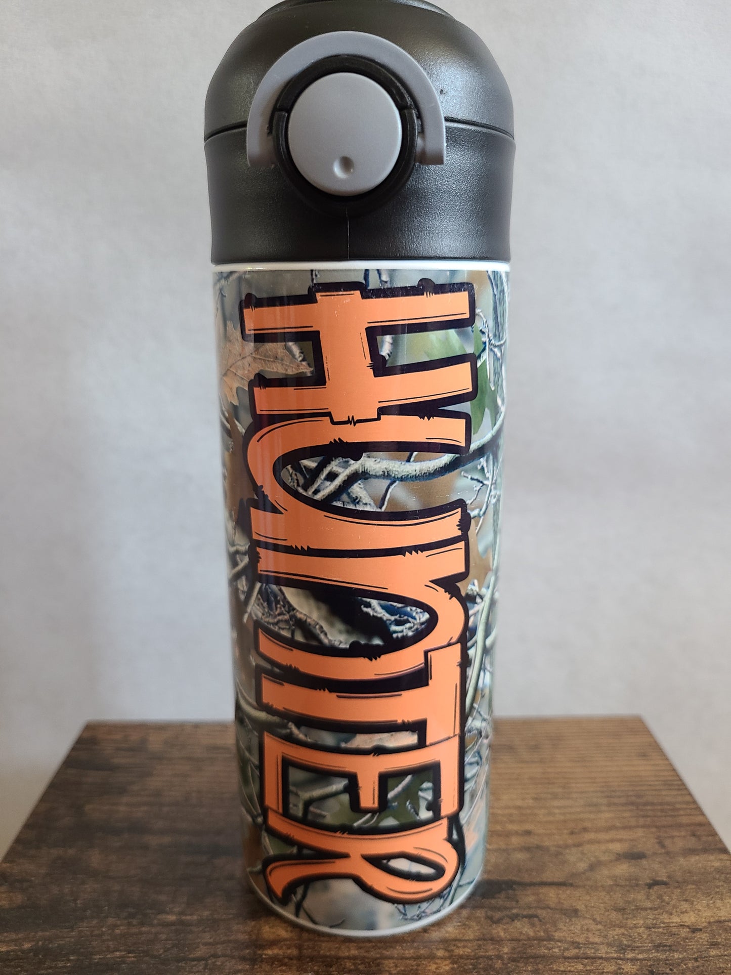 12 oz water bottle with a black lid that has a built-in straw, locking push button and handle. This design features real tree camo with orange personalization. Back to School.
