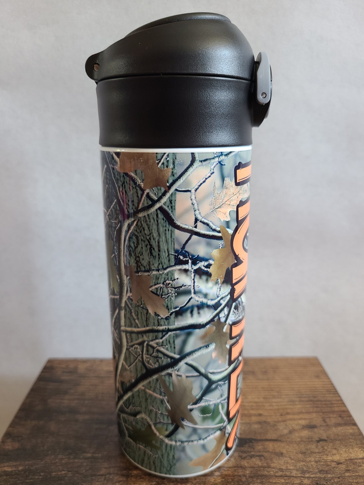 12 oz water bottle with a black lid that has a built-in straw, locking push button and handle. This design features real tree camo with orange personalization. Back to School.