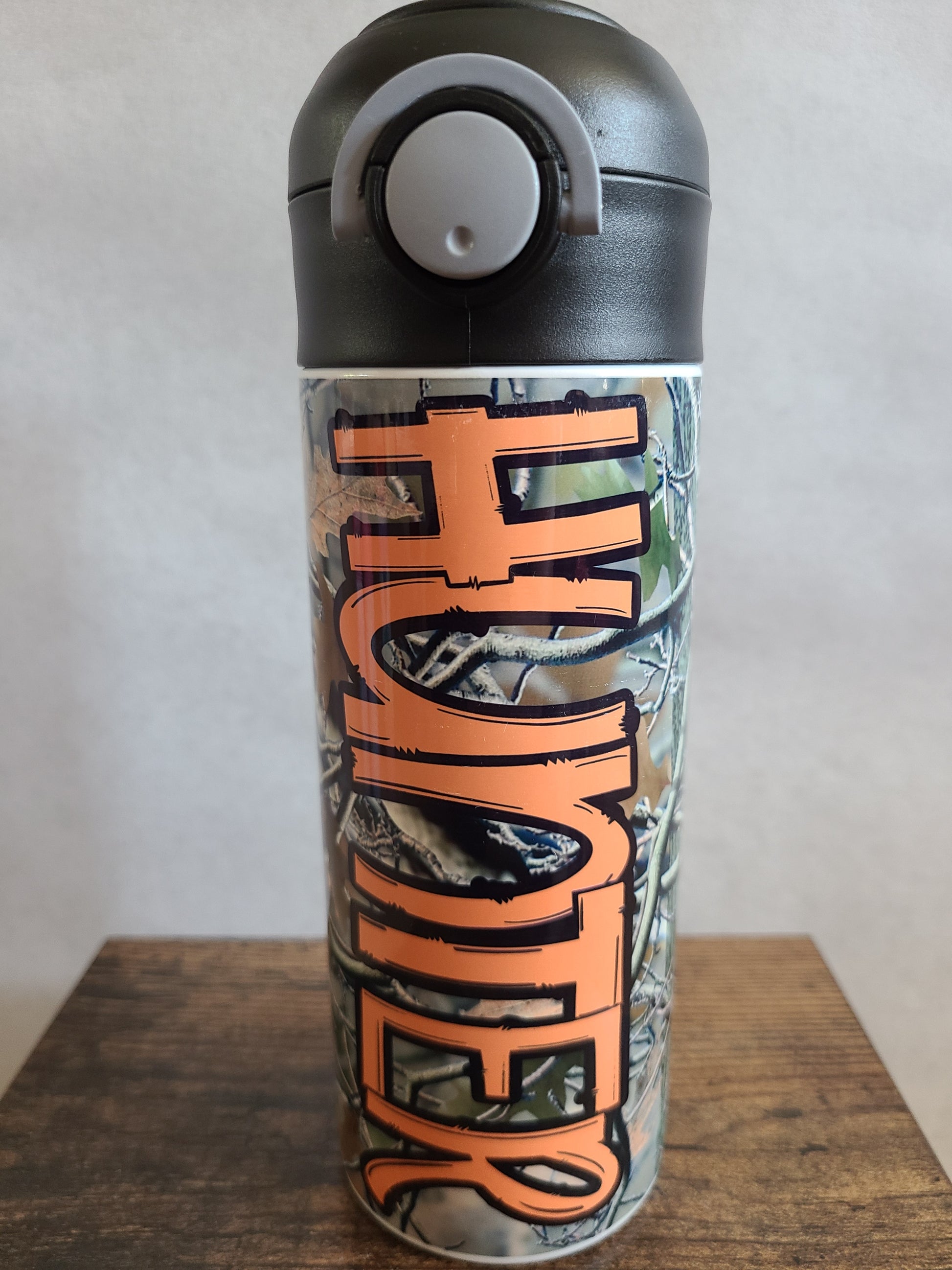 12 oz water bottle with a black lid that has a built-in straw, locking push button and handle. This design features real tree camo with orange personalization. Back to School.