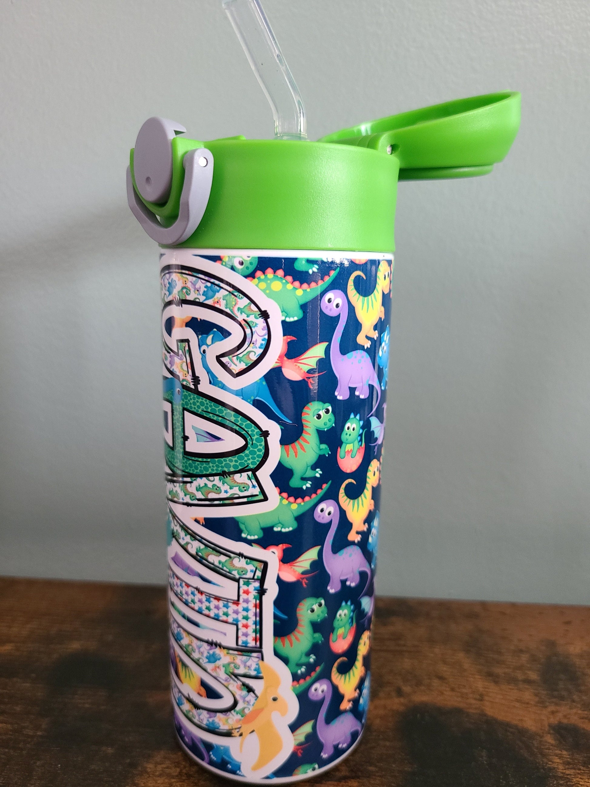 12 oz water bottle with flip top and built in straw. Colorful dinosaur themed water bottle for kids. Back to school water bottle for kids. Dinosaurs in yellow, green, purple and blue with navy blue background and green lid.