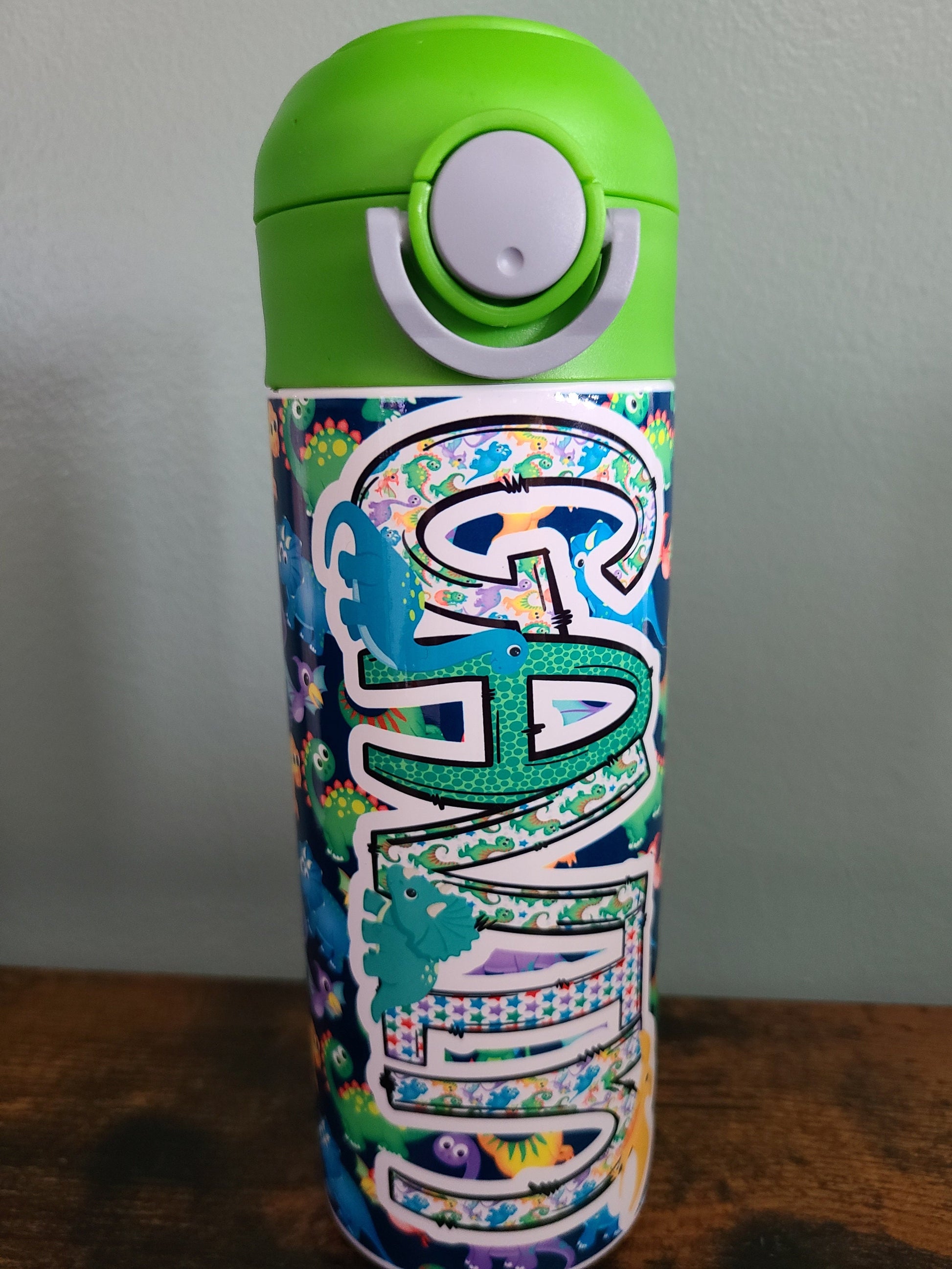 12 oz water bottle with flip top and built in straw. Colorful dinosaur themed water bottle for kids. Back to school water bottle for kids. Dinosaurs in yellow, green, purple and blue with navy blue background and green lid.