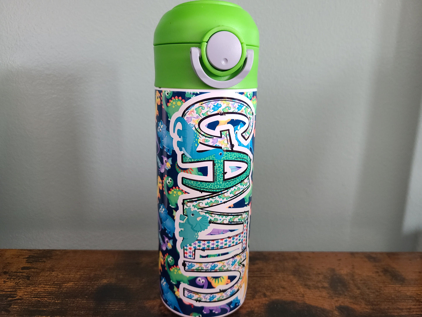 12 oz water bottle with flip top and built in straw. Colorful dinosaur themed water bottle for kids. Back to school water bottle for kids. Dinosaurs in yellow, green, purple and blue with navy blue background and green lid.