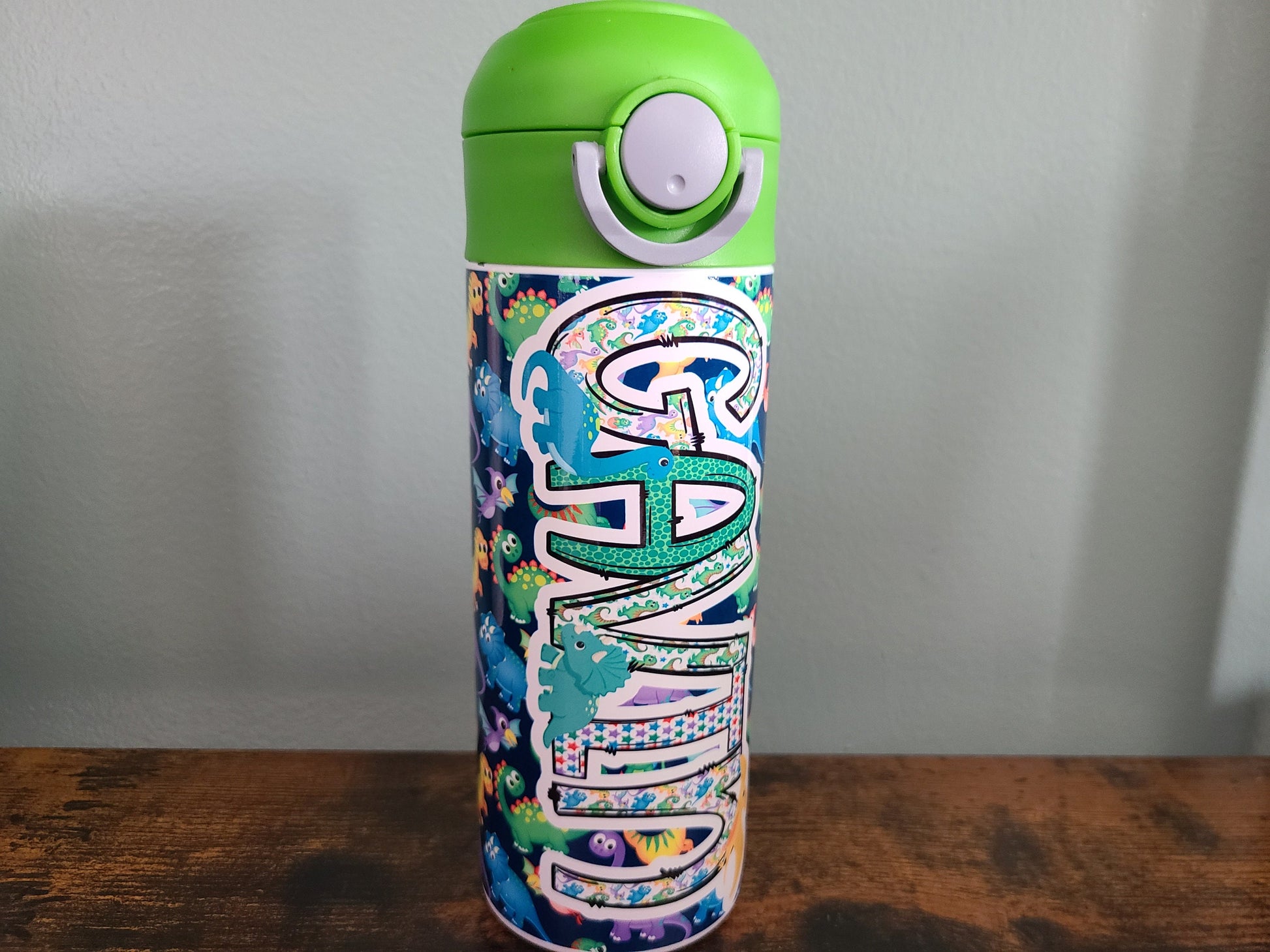 12 oz water bottle with flip top and built in straw. Colorful dinosaur themed water bottle for kids. Back to school water bottle for kids. Dinosaurs in yellow, green, purple and blue with navy blue background and green lid.