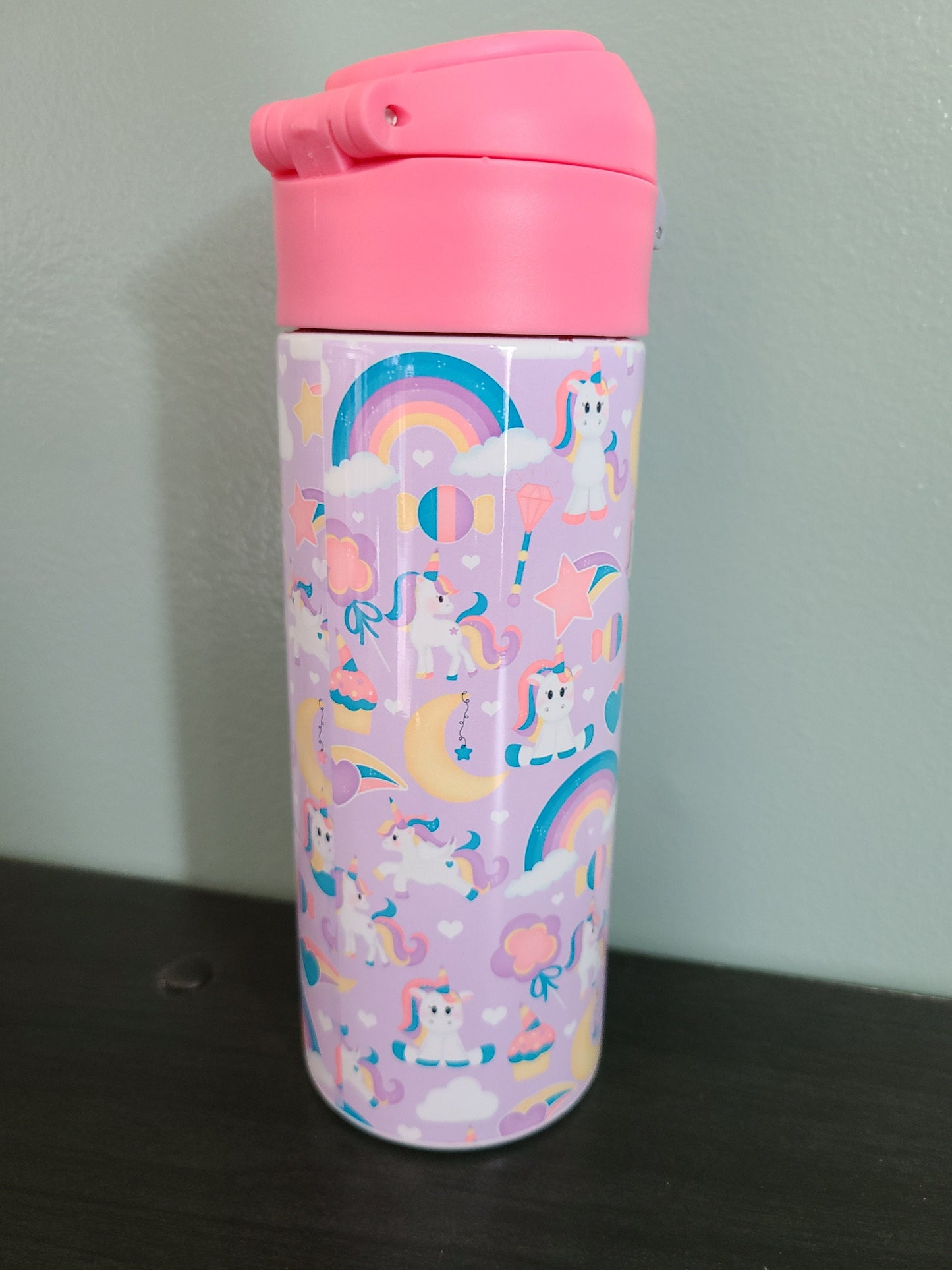 12 oz water bottle with flip top and built in straw. Unicorn themed water bottle for kids. Back to school water bottle for kids. Light purple background with white unicorns that have colorful hair, moons, stars and rainbows with a pink lid.