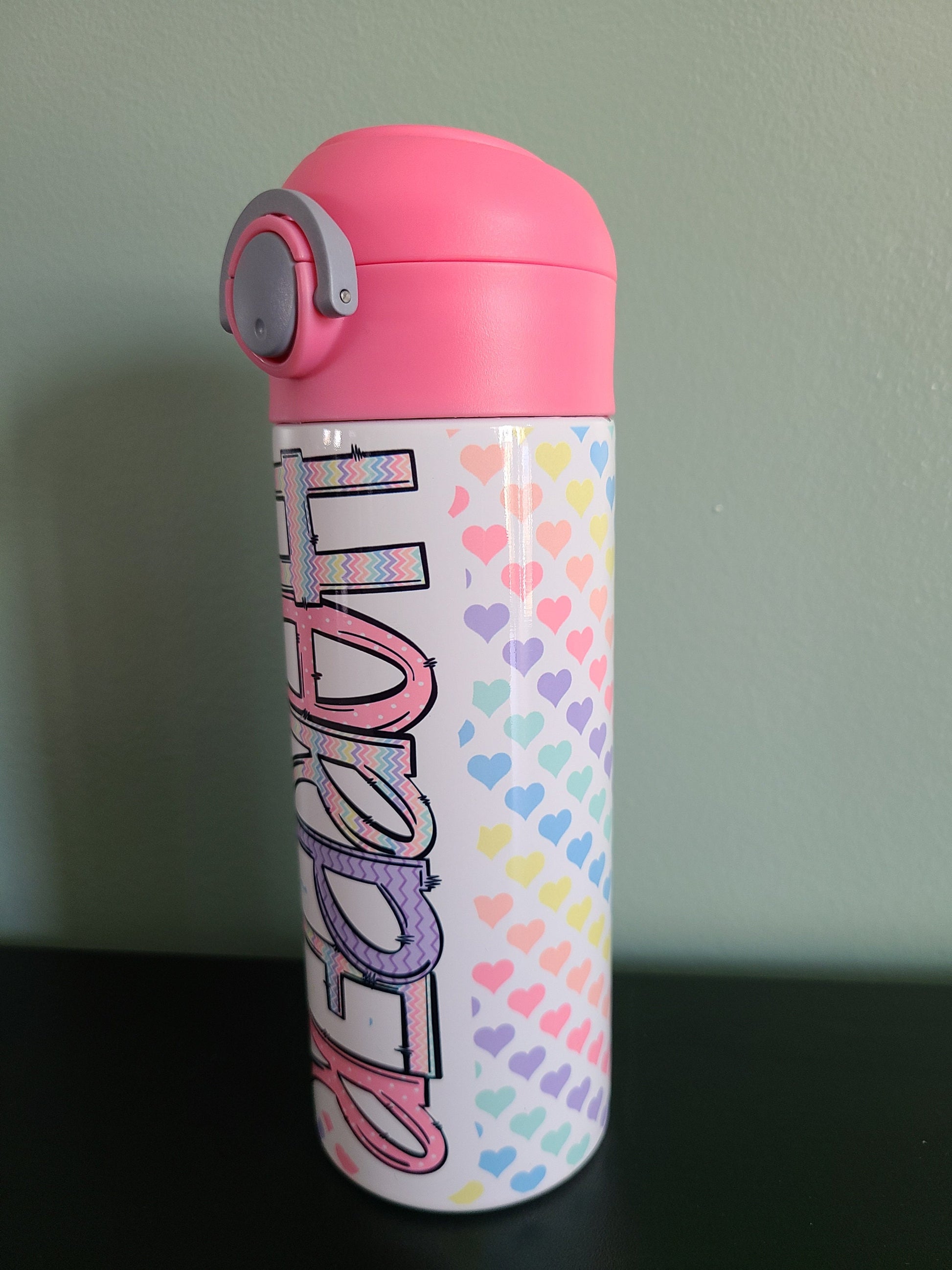 12 oz water bottle with flip top and built in straw. Pastel colored heart themed water bottle for kids. Back to school water bottle for kids. Hearts in pastel colors with a complimentary design personalization and pink lid.