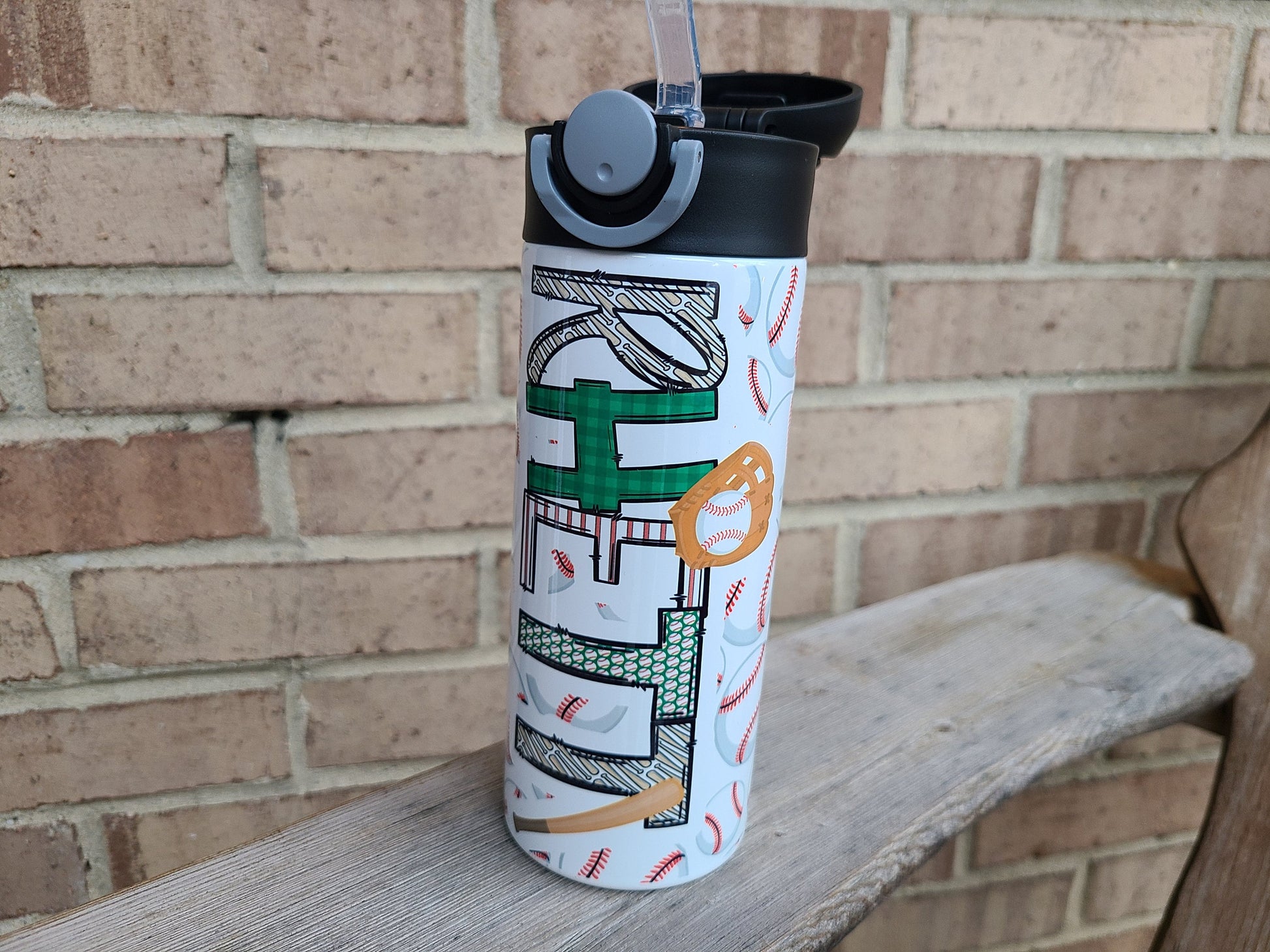 12 oz water bottle with a black lid that has a built-in straw, locking push button and handle. This design features multiple baseballs with a glove and bat print. Personalized in coordinating colors. Back to School. Lid pops open to show silicone straw.