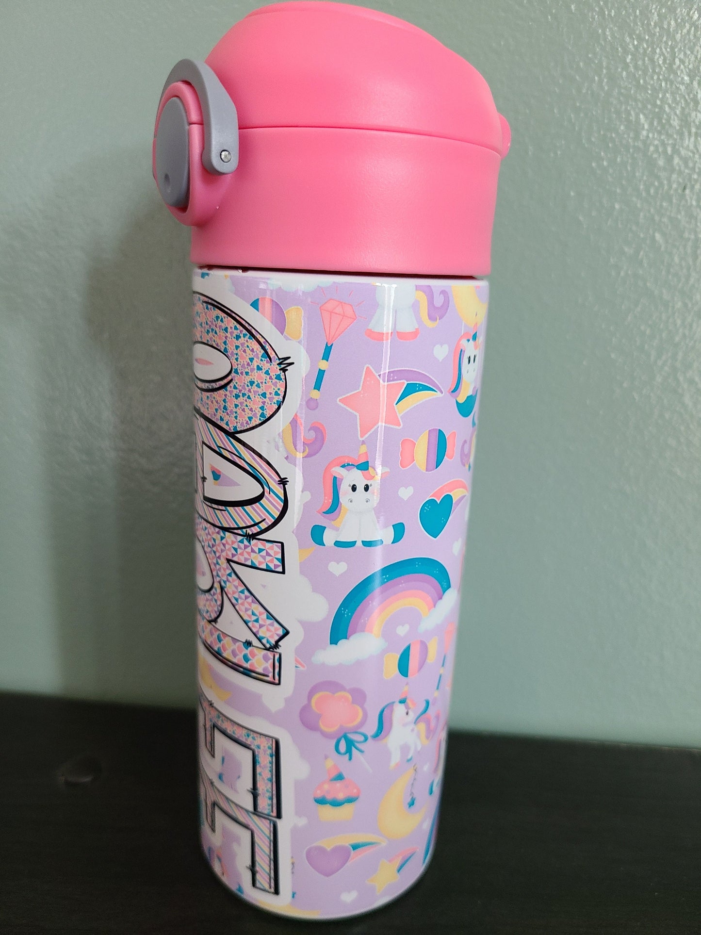 12 oz water bottle with flip top and built in straw. Unicorn themed water bottle for kids. Back to school water bottle for kids. Light purple background with white unicorns that have colorful hair, moons, stars and rainbows with a pink lid.