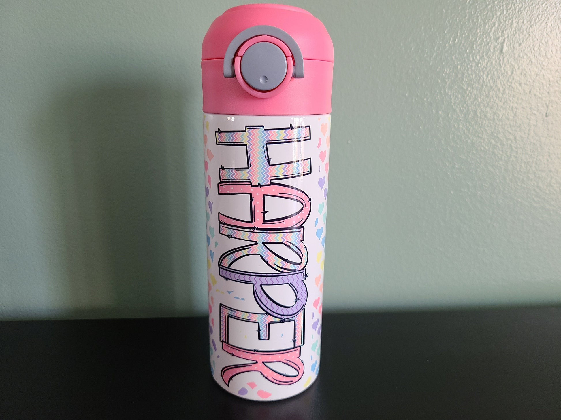 12 oz water bottle with flip top and built in straw. Pastel colored heart themed water bottle for kids. Back to school water bottle for kids. Hearts in pastel colors with a complimentary design personalization and pink lid.