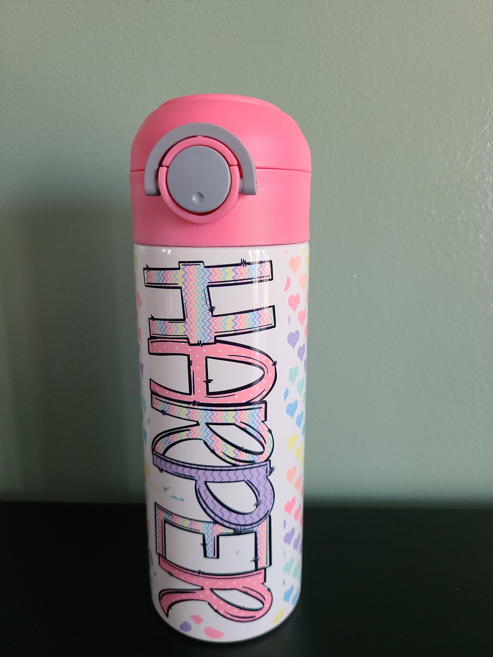 12 oz water bottle with flip top and built in straw. Pastel colored heart themed water bottle for kids. Back to school water bottle for kids. Hearts in pastel colors with a complimentary design personalization and pink lid.